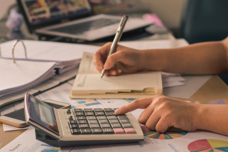 Mãos anotando lançamentos ao lado de uma calculadora, sobre documentos contábeis